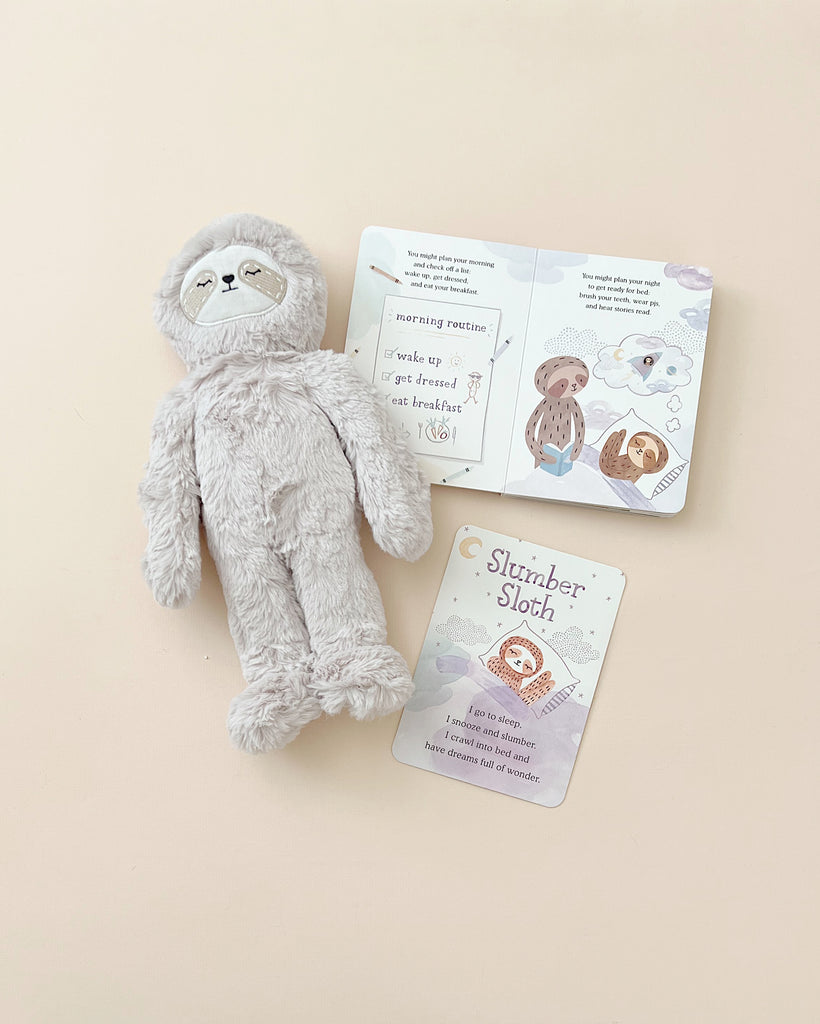 A Slumberkins Sloth Kin + Lesson Book - Routines lying next to an open children's book about sloths, displayed on a light beige background.