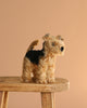 A Steiff Mohair Terri Welsh Terrier plush toy, 10 inches tall in brown and black, stands on a wooden stool against a plain light brown background.