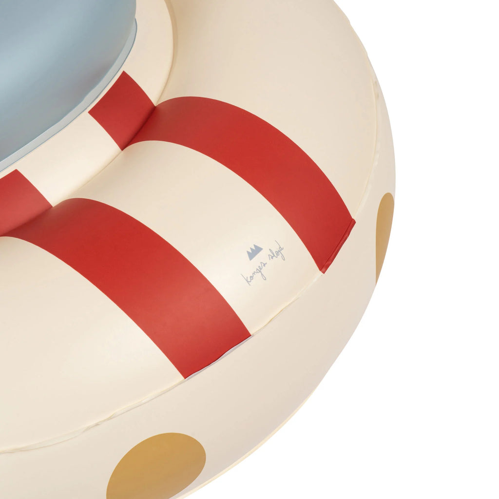 A close-up view of a beige Inflatable Baby Swim Ring - Car for 1-2 years with thick red stripes and brown polka dots. Crafted from durable PVC, the logo "konges slojd" is visible on the side.