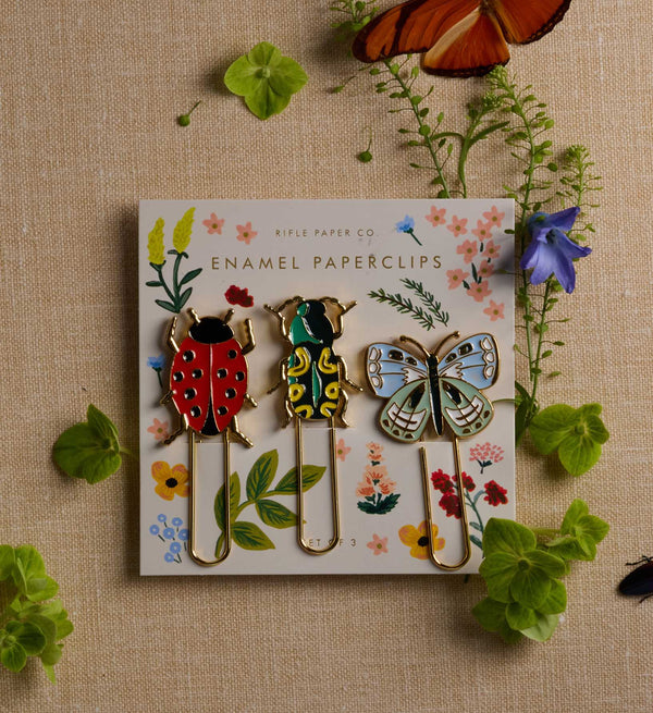 The Curio Enamel Paper Clip Set features three paperclips shaped like a ladybug, beetle, and butterfly, each with colorful enamel detailing and displayed on a floral card. Surrounded by green plants, a purple flower, and two orange butterfly wings on a beige background, this delightful set has an overall product size of 4 x 4 x 0.5 inches.