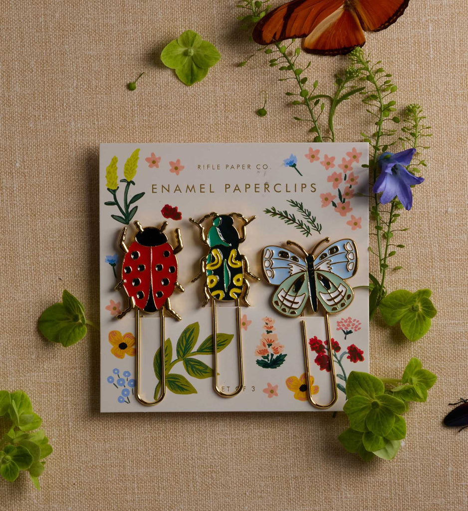 The Curio Enamel Paper Clip Set features three paperclips shaped like a ladybug, beetle, and butterfly, each with colorful enamel detailing and displayed on a floral card. Surrounded by green plants, a purple flower, and two orange butterfly wings on a beige background, this delightful set has an overall product size of 4 x 4 x 0.5 inches.