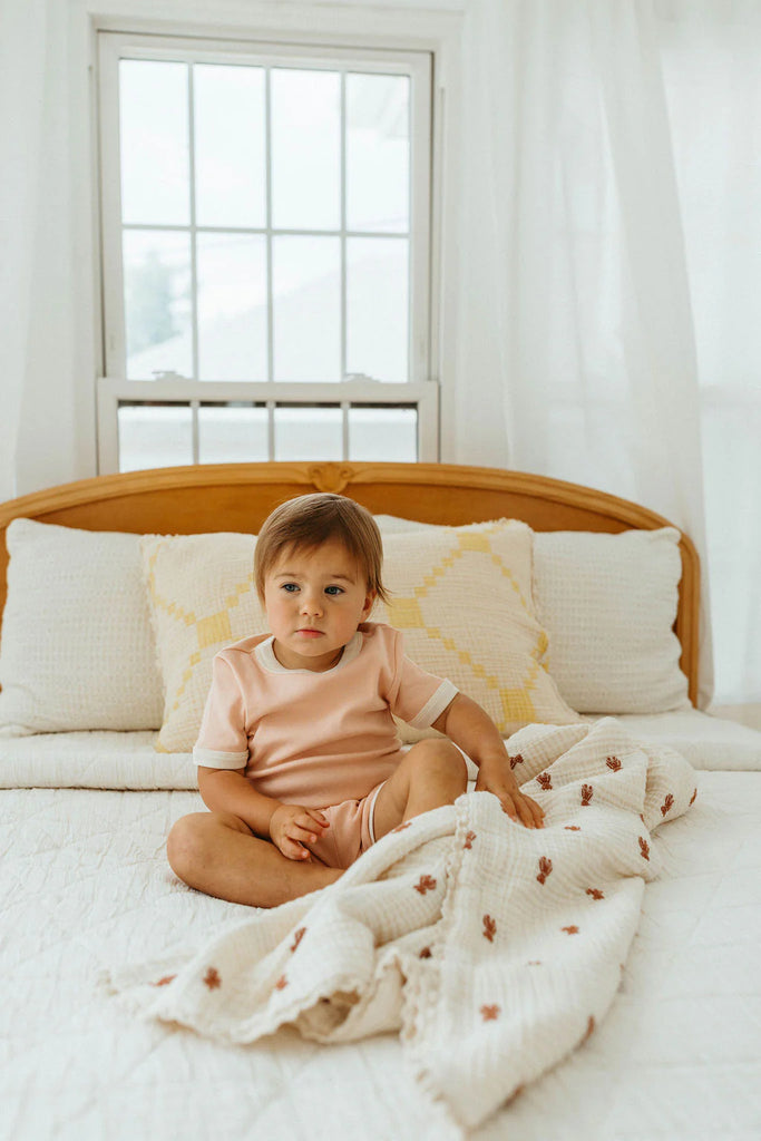 A young child in a light pink outfit sits cross-legged on a bed with the Organic Cotton Layered Gauze Baby Blanket - Teddy (ships in one week), against a wooden headboard and a large window with white curtains.