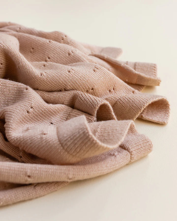 A close-up image of a crumpled beige Handmade Merino Wool Bibi Blanket - Apricot with a textured, knitted pattern and small holes distributed throughout the fabric. The background is light-colored, emphasizing the blanket's details.
