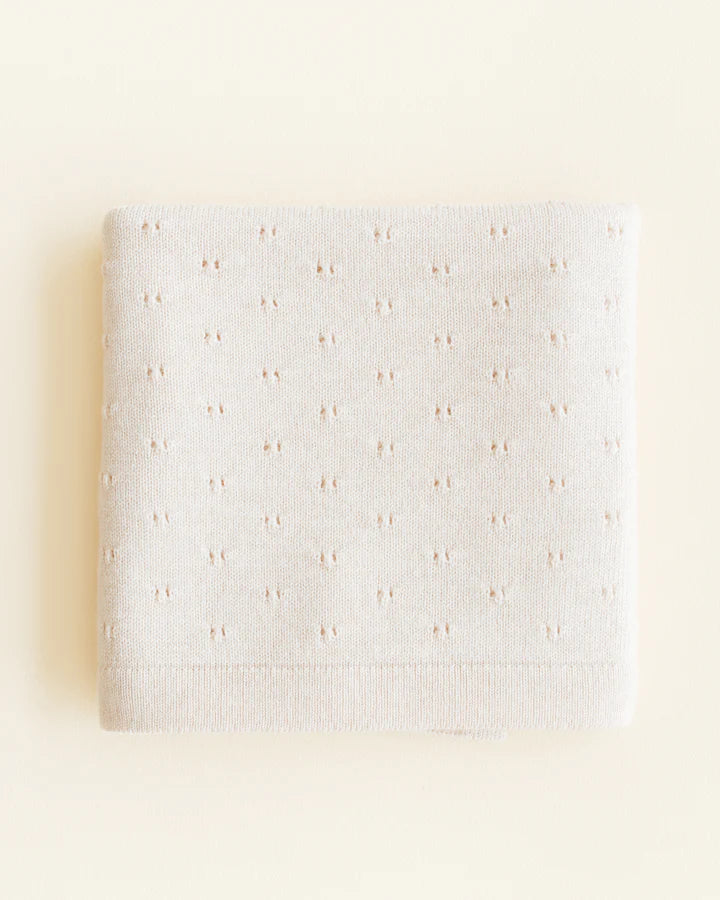 A folded Handmade Merino Wool Bibi Blanket in Cream with a knit pattern that includes small, evenly spaced pink rosettes, presented against an off-white background.