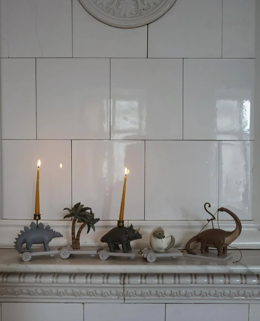 Four whimsical, Dinosaur Birthday Train With Beeswax Candles displayed on a white mantelpiece: a dinosaur, a palm tree, a snail, and an elephant, each holding a lit candle, against a tiled