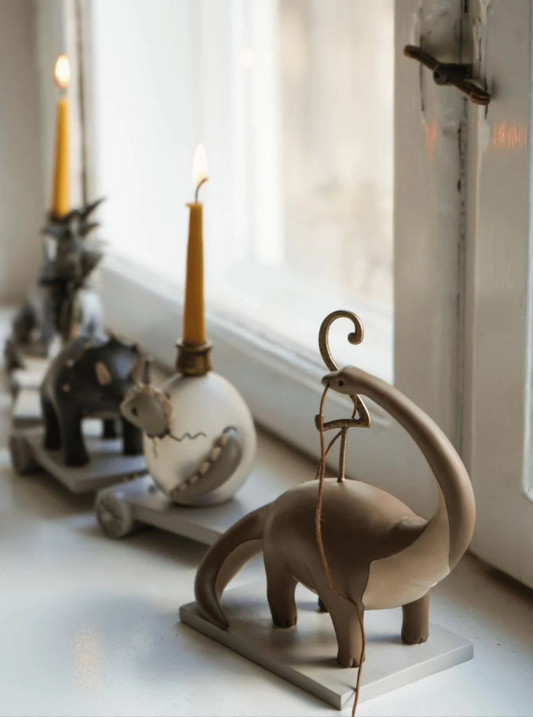 A whimsical display featuring a wooden elephant sculpture with a lit candle on its back, beside a window with two other candle holders, one shaped like an elephant and another like the Dinosaur Birthday Train With Beeswax Candles.