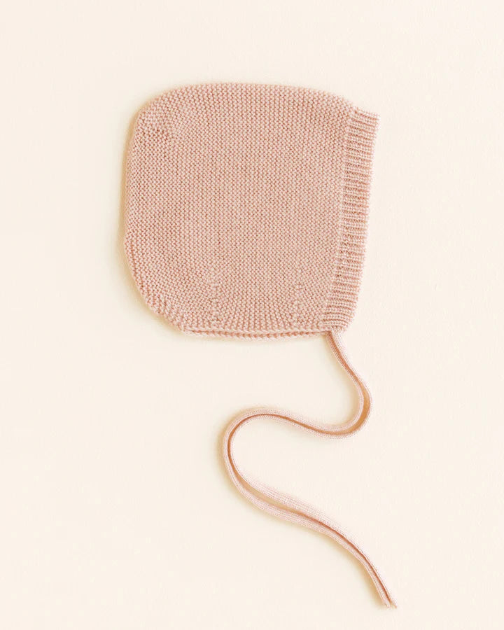 A light pink, seamless knitted Handmade Merino Wool Newborn Bonnet - Apricot with a delicate ribbed texture and two thin, long strings, placed neatly against a soft beige background.