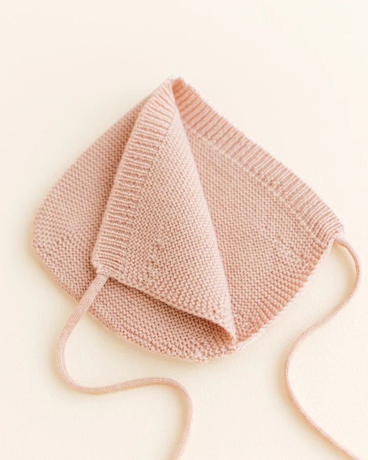 A pale pink Handmade Merino Wool Newborn Bonnet - Apricot placed on a light beige background, displayed unfolded with its triangular silhouette evident and strings on the side.