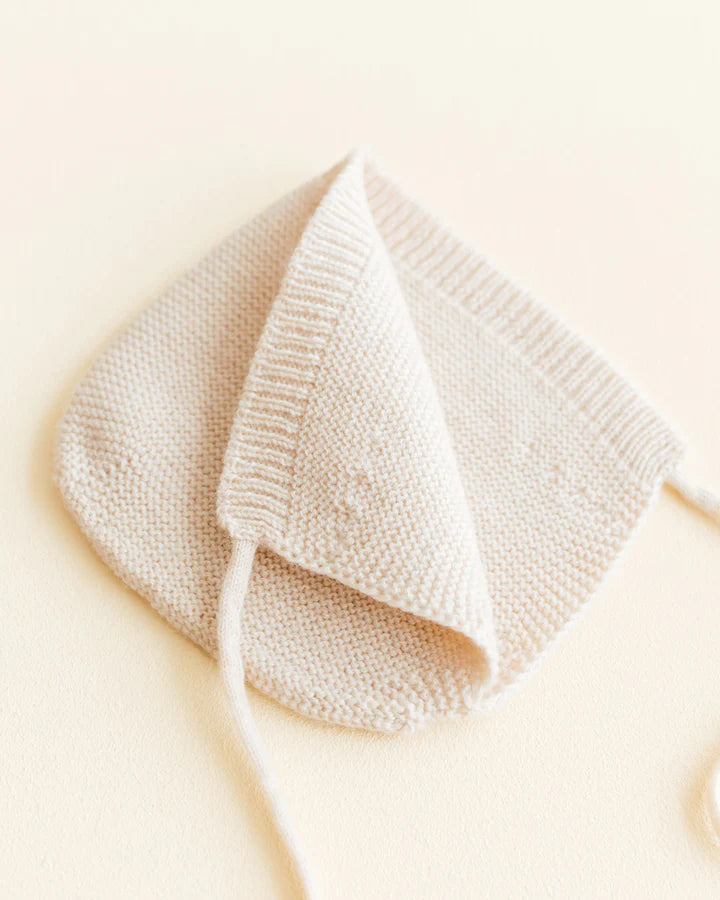 A hand-knit, Cream-colored Handmade Merino Wool Newborn Bonnet with a delicate texture, displayed on a light beige background. The bonnet features a ribbed hem and thin ties.