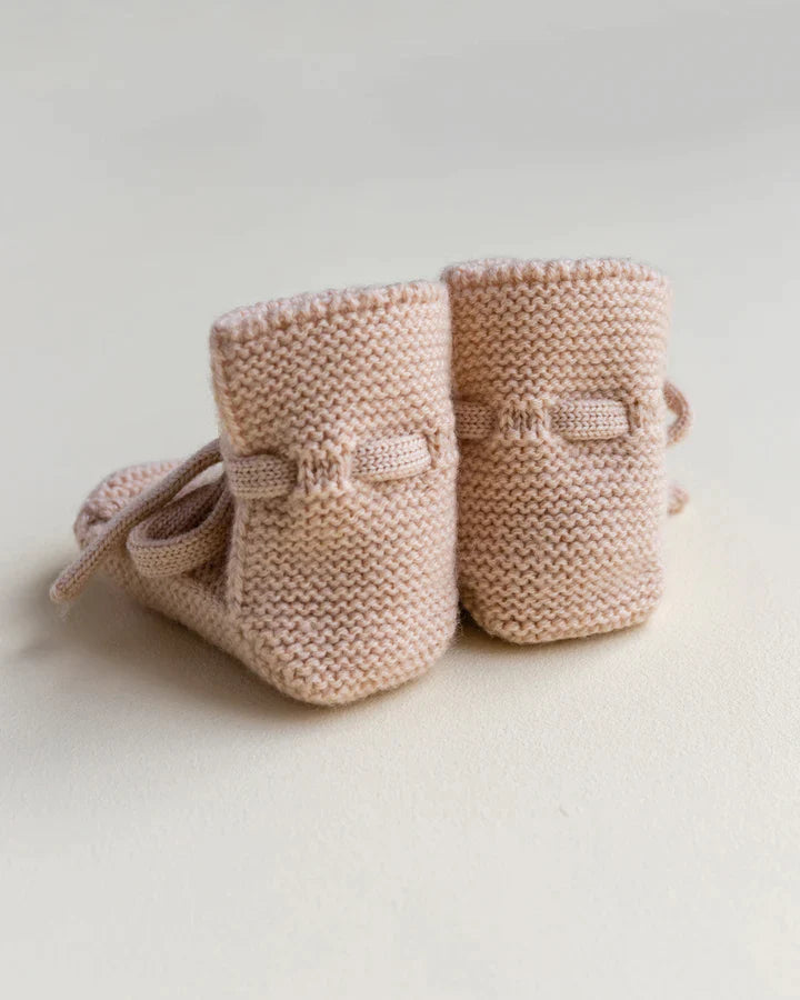 A pair of hand-knitted, soft pink apricot merino wool booties on a light beige surface, showing intricate stitching and tied with delicate laces.