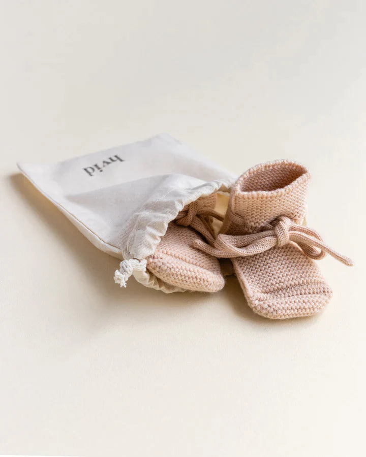 A pair of small, apricot merino wool baby booties with laces, placed beside a white cloth bag labeled “plan” on a soft beige background.