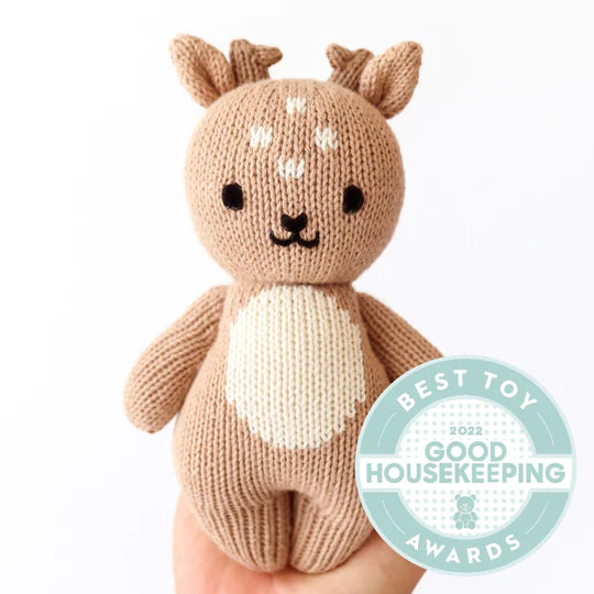 A small Cuddle + Kind Baby Fawn hand-knit plush toy deer with a tan body, white belly, and antlers being held in a person's hand, crafted from Peruvian cotton yarn, with a "best toy