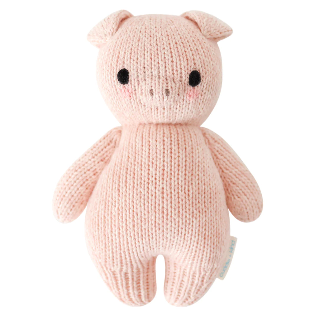 A Cuddle + Kind Baby Piglet plush toy, with a pale pink hand-knit body, black button eyes, and small ears, standing upright against a white background.