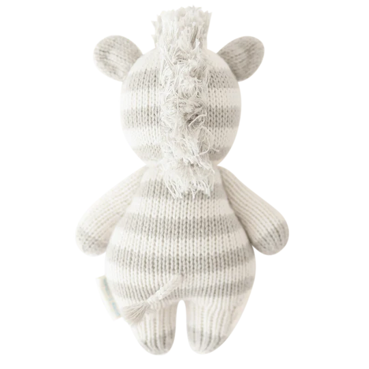 A hand-knit Cuddle + Kind Baby Zebra toy in a white and light grey color scheme, viewed from the back, featuring distinct striped patterns and a fluffy mane.
