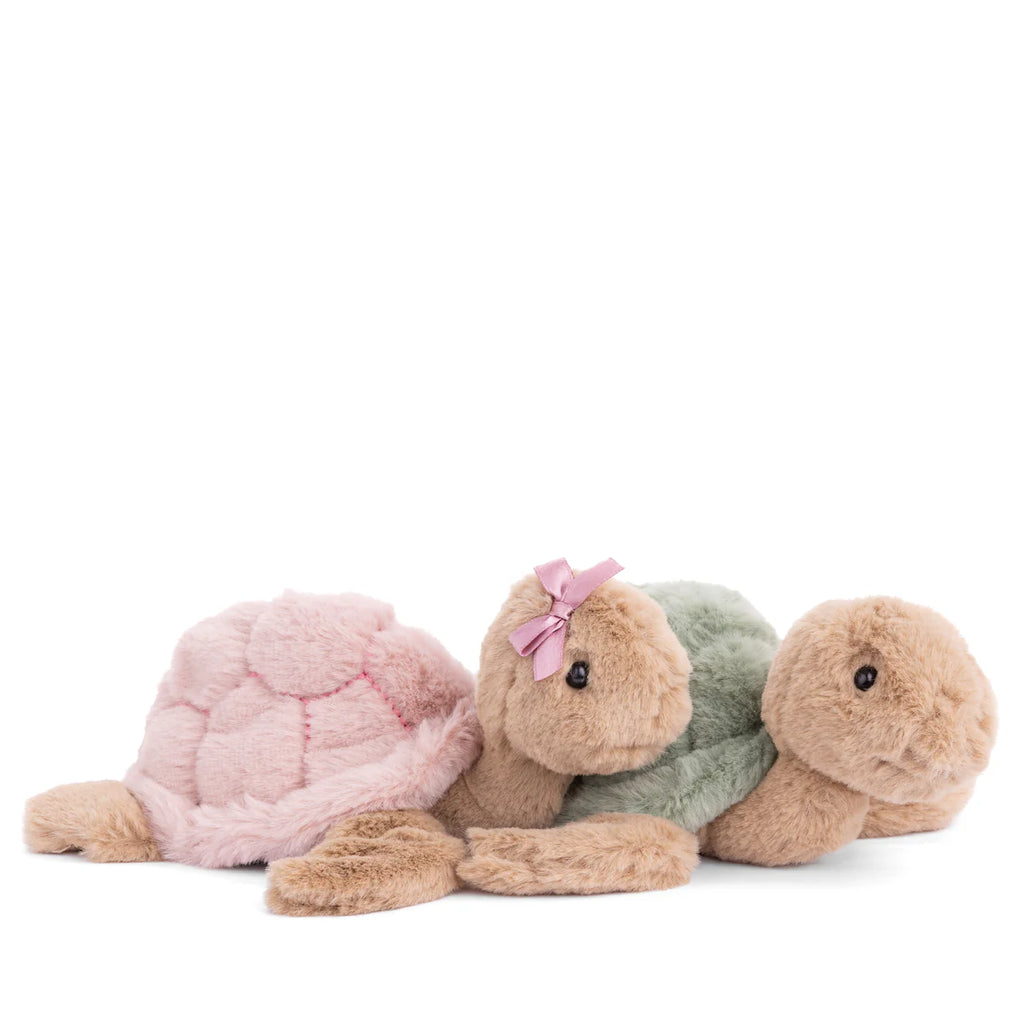 Two Bukowski Baby Sandy Sea Turtle plush toys are side by side: one has a green shell, and the other a pink shell with a pink bow. Both feature soft brown bodies and black button eyes.