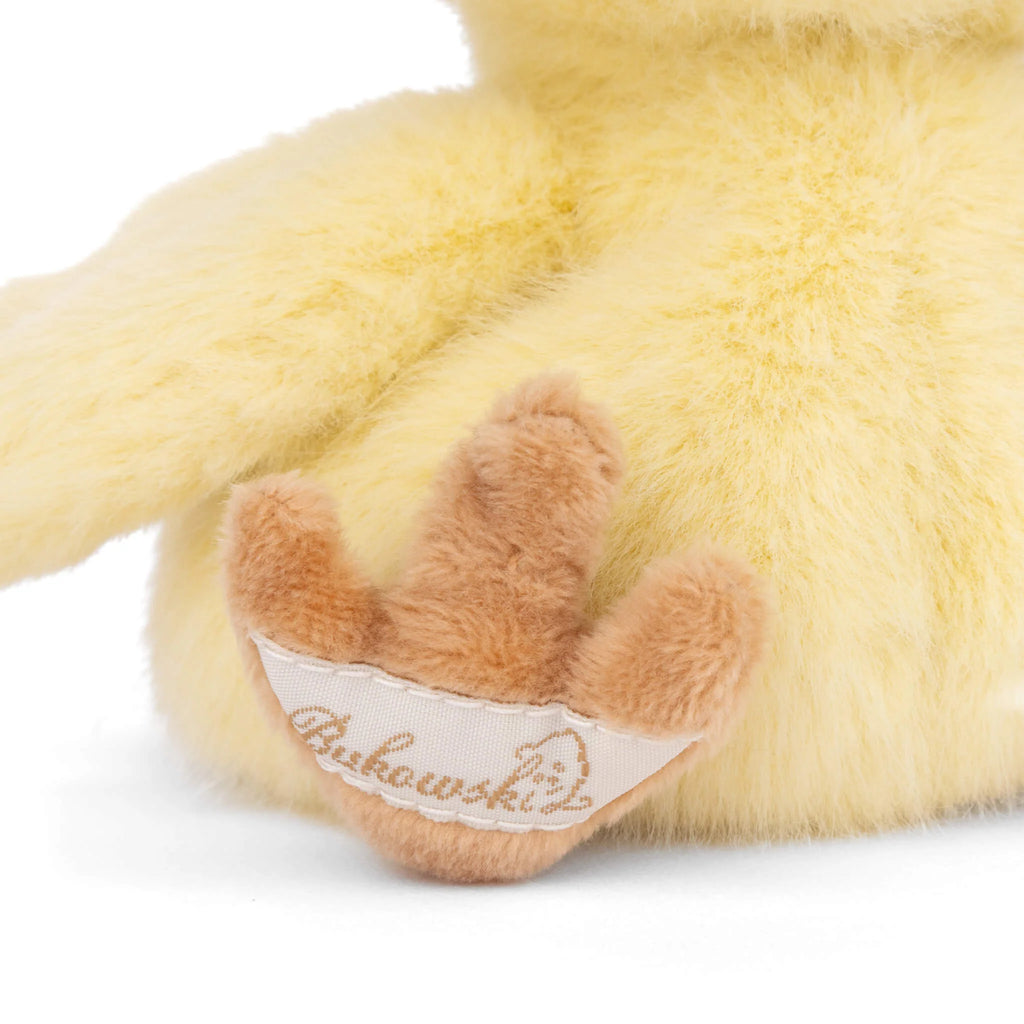 Close-up of Bukowski - Baby Chicky’s soft yellow plush paw, featuring brown toes and a white fabric tag reading "Bukowski" in script.