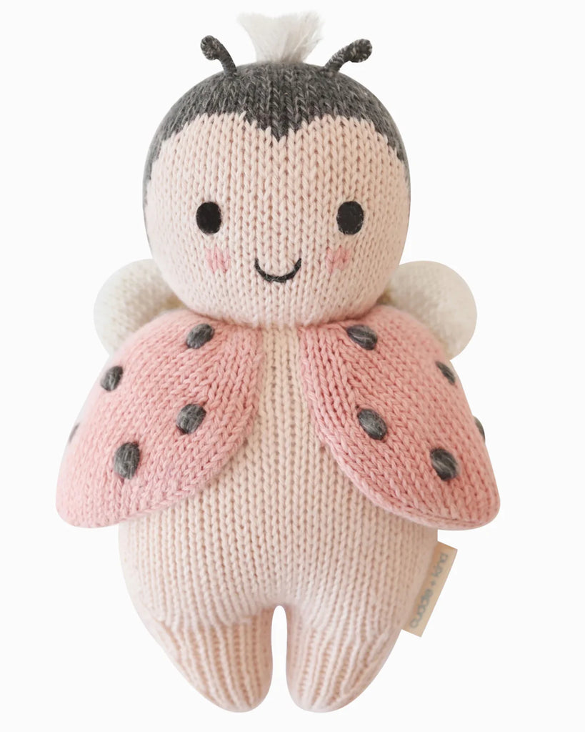 The Cuddle + Kind Baby Ladybug is a hand-knit plush toy made from natural Peruvian cotton yarn, featuring pink wings, black spots, and blushing cheeks. Crafted using fair trade practices for a soft and adorable look.