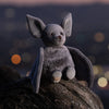 The Bukowski - Baltazar Bat, a gray plush bat with large ears, sits on a tree trunk at dusk, its silhouette softly framed by blurred city lights in the background.