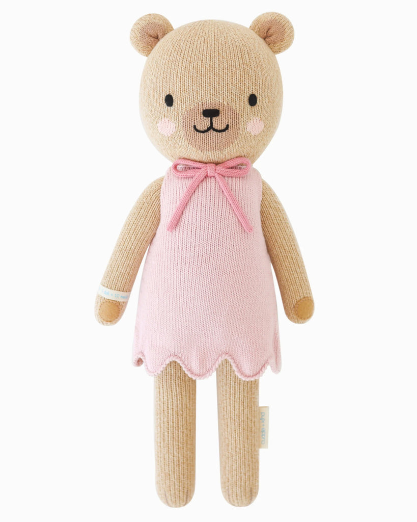 Cuddle + Kind Bella the Honey Bear is a hand-knit doll with light brown fur, a pink dress with scalloped hem, and a pink bow at the neck—smiling sweetly and ready for cuddles on a white background.