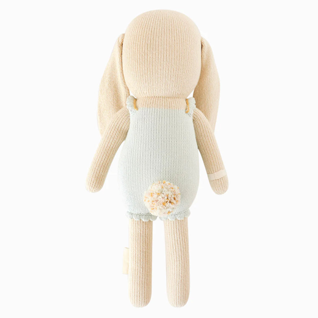 The Cuddle + Kind Briar the Bunny - Light Blue is a hand-knit, 100% cotton bunny doll from Peru, featuring long floppy ears, a fluffy pom-pom tail, and a light blue romper, shown from the back.