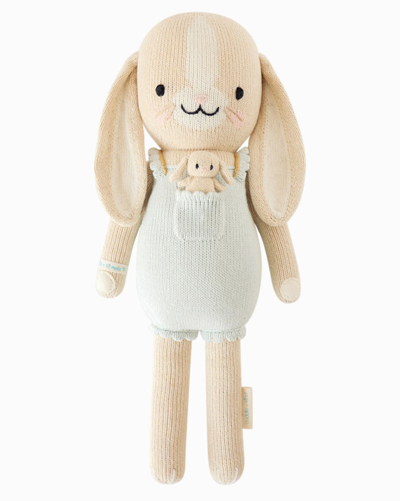 Cuddle + Kind Briar the Bunny - Light Blue is a beige, hand-knit doll made from premium cotton yarn, featuring long ears, embroidered eyes and smile, pink cheeks, and a light blue romper with a cute bunny face—handmade in Peru.