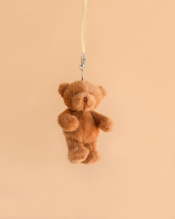 A small brown Bukowski - Happy Mavi Bag Charm XL plush keychain is hanging by a metal clasp and a light-colored strap against a plain beige background, making it a perfect giftable stuffed animal for any occasion.