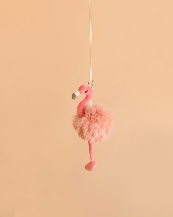 The Bukowski - Phoebe Flamingo Bag Charm, a fluffy pink plush flamingo keychain with a yellow ribbon, hangs against a plain light peach background—making it the perfect adorable accessory for your bag or keys.