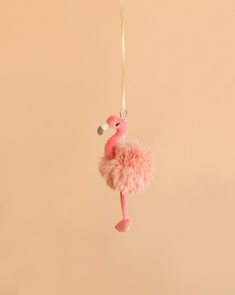 The Bukowski - Phoebe Flamingo Bag Charm, a fluffy pink plush flamingo keychain with a yellow ribbon, hangs against a plain light peach background—making it the perfect adorable accessory for your bag or keys.