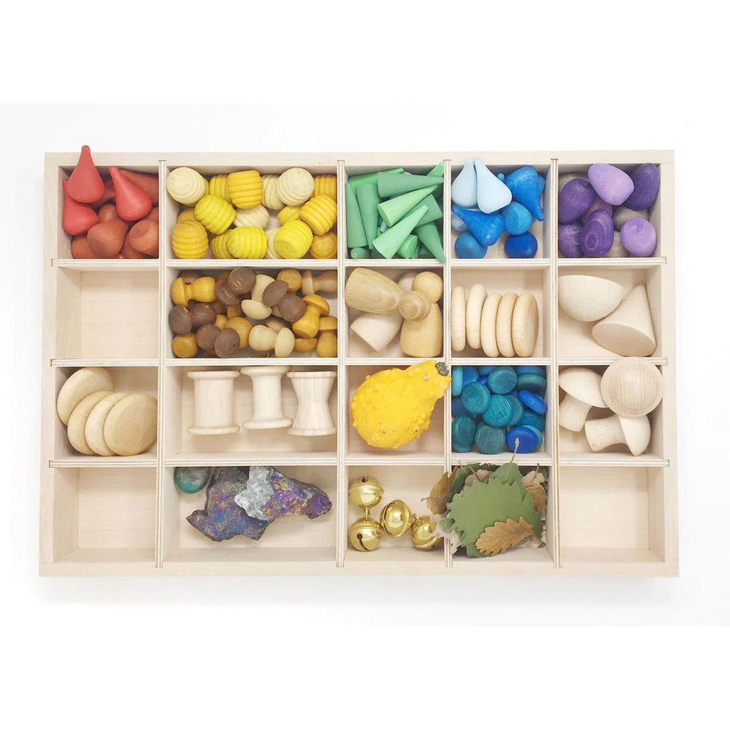 The Grapat Tinker Tray is a sustainable wooden organizer with 18 compartments, ideal for displaying small, colorful objects—cones, cylinders, pebbles—for loose-part play and sorting by color and shape in a rainbow sequence.