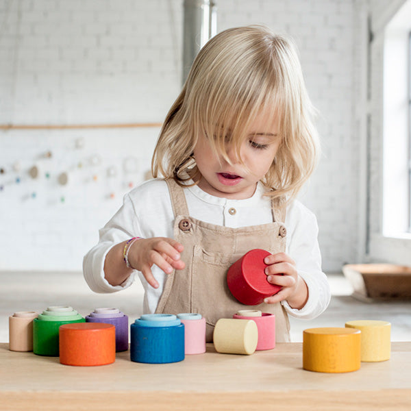 A young child with light hair playing with Grapat Nesting Bowls on a table in a bright room with a white brick wall backdrop.