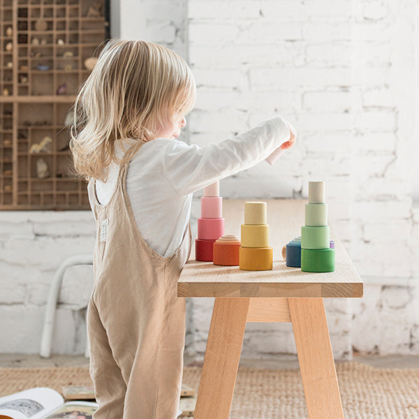 A young child interacts with Grapat Nesting Bowls on a wooden table in a brightly lit room with a white brick background.