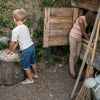 Two children, a boy and a girl, play outside a rustic wooden hut. The boy stands holding a Grapat Small Storage Box while the girl peeks inside the hut. Outdoor play setting.
