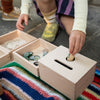 A child sits on a colorful rug, putting wooden caps into a small slot on a Grapat Permanence Box, with other open boxes containing bottle caps and rings beside them.