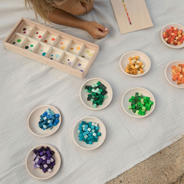 A child playing with Grapat Mix & Match colorful wooden beads and a wooden sorting box on a light fabric surface outdoors. Wooden bowls contain organized beads by color.