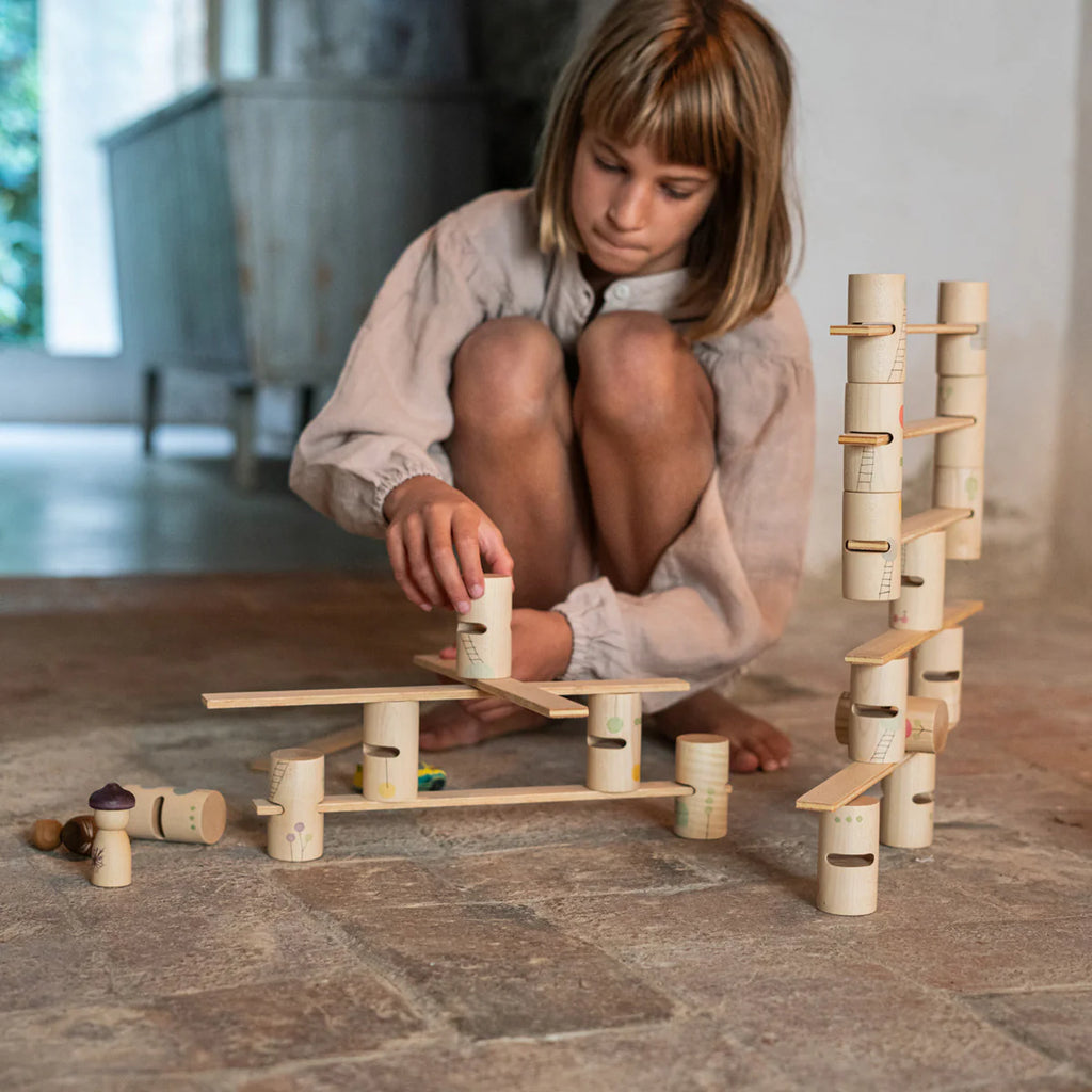 A child with short hair kneels on the tiled floor, absorbed in creating a marble run using the Grapat Woodland Building Set. The unfinished wooden structure shines in the sunlight streaming through a window.