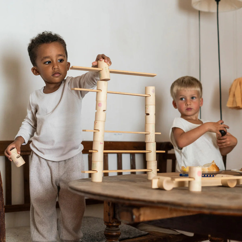 Two young children enjoy endless play with the Grapat Woodland Building Set indoors. One carefully places blocks on a tall structure while the other holds a toy in the background, both near a wooden table and lamp in their cozy room. Ships in approximately one week.