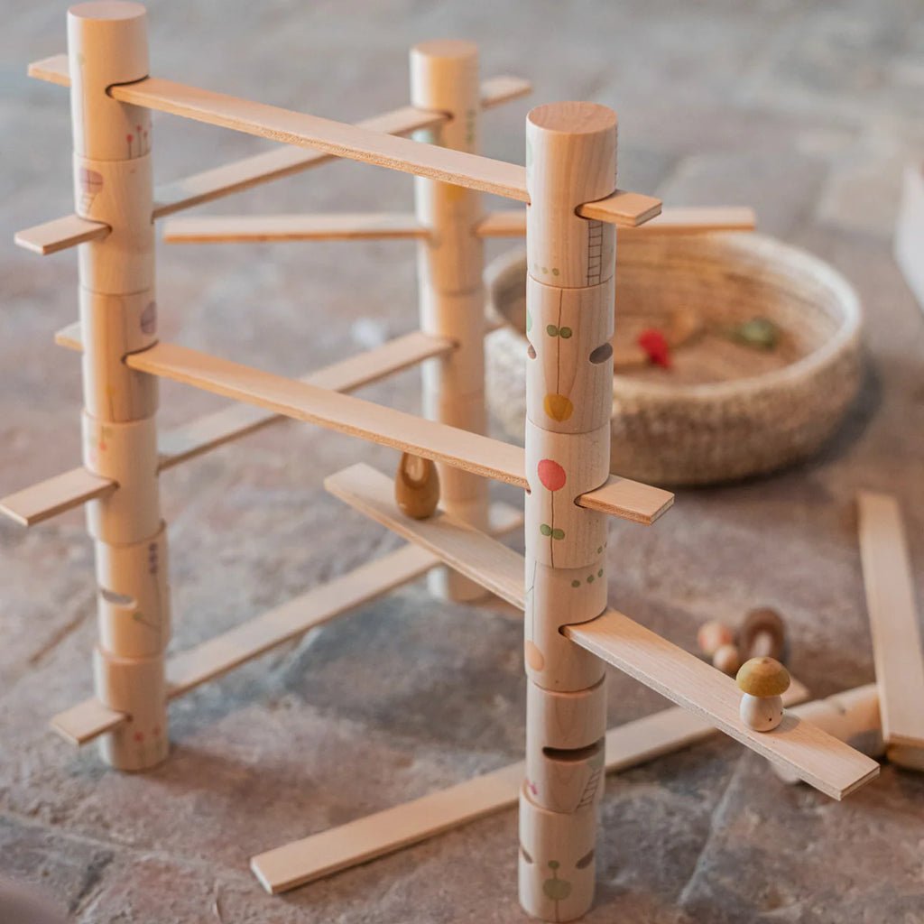 The Grapat Woodland Building Set, made of natural wood with colorful patterns, is assembled on the floor. Wooden marbles are prepared to roll down this engaging set, promising endless fun. A woven basket and additional pieces can be seen in the background. Ships in approximately one week.