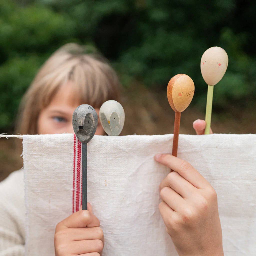 A child displays a white cloth with Grapat Twin Souls Puppets—four wooden spoon puppets each adorned with simple faces. The lush green backdrop enhances these storytelling tools, igniting creativity. Ships in approximately one week.