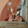 Pairs of hands hold Grapat Twin Souls Puppets with painted faces behind a rustic window pane, weaving a storytelling charm. An orange gingham cloth is partially visible in the background, enhancing the nostalgic allure of children's toys. Ships in approximately one week.