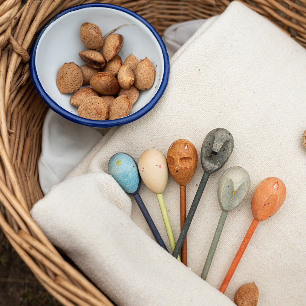 A wicker basket with a white cloth holds a blue-rimmed bowl of shelled almonds. Beside it, six colorful Grapat Twin Souls Puppets-inspired wooden spoons with carved double faces invite imagination and storytelling.