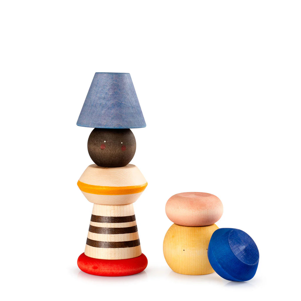 The Grapat Serendipity Stacking Toy features a handcrafted wooden figure with a blue cone hat, round head, striped body, and red base. It includes two blue pieces and one peach piece on light wood, ideal for children who love building stacking towers. Ships in approximately one week.