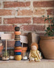 A small doll in a yellow dress sits on a shelf beside the Grapat Serendipity Stacking Toy and colorful wooden toys. Nearby, potted plants and sprouting jars rest against a rustic brick wall.