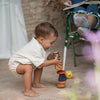 A toddler squats stacking vibrant rings from the Grapat Serendipity Stacking Toy, while an adult sits with a book in a cozy, softly lit rustic setting. (Ships in approximately one week.).