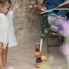 A toddler stretches their arms to the colorful, handcrafted Grapat Serendipity Stacking Toy on the ground, smiling. Nearby, a blurred adult sits in a green chair with a book, legs crossed and feet up. An apple lies next to the 8-piece stacking tower.