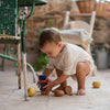 A toddler crouches on a tiled patio, using the Grapat Serendipity Stacking Toy to build a colorful tower. Dressed in light clothing, the child is surrounded by rustic furniture, with a wicker basket and lush plants enhancing the handcrafted ambiance.