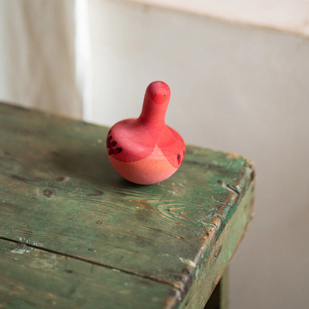 A small, pink Grapat Chill Bird Rocking Toy with a handle rests on an aged green wooden surface with black patterns near the table's edge, poised as if to fly. A beige wall forms the backdrop.