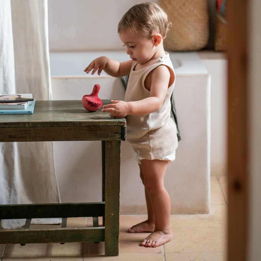 A toddler stands barefoot beside a wooden table, playing with the Grapat Chill Pink Bird Rocking Toy. The child wears a light sleeveless outfit, surrounded by neutral decor and soft lighting that creates a calming atmosphere.