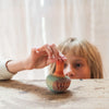 A child with blonde hair peeks over a wooden table, gently holding a small Grapat Mellow Orange Bird Rocking Toy. Its playful wings appear ready to take flight against the neutral curtains that provide a calming backdrop.