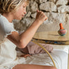 A child on a couch, in a white shirt, plays with a Grapat Mellow Orange Bird Rocking Toy on a rustic table. The setting is completed by a textured stone wall decorated with vibrant feathers.