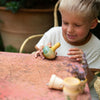 A young child with short blonde hair smiles while playing with a small wooden toy on a weathered red table outdoors. Nearby, a Grapat Flowing Yellow Bird Rocking Toy flits around, catching the sunlight. Small flower pots adorn the table, and a potted plant sits in the background.